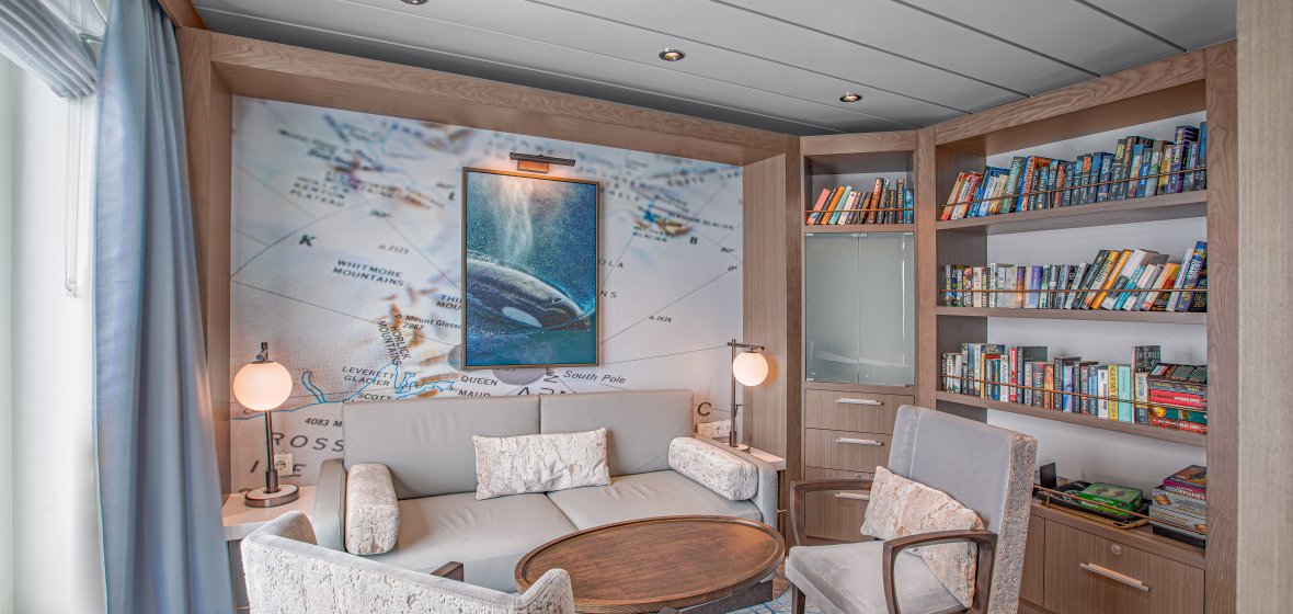 A contemporary cruise ship cabin featuring twin beds with cream-colored bedding, purple and coral accent pillows, and a neutral color palette. The room showcases modern furnishings including a padded headboard, ambient lighting, and upscale décor typical of premium cruise line accommodations.