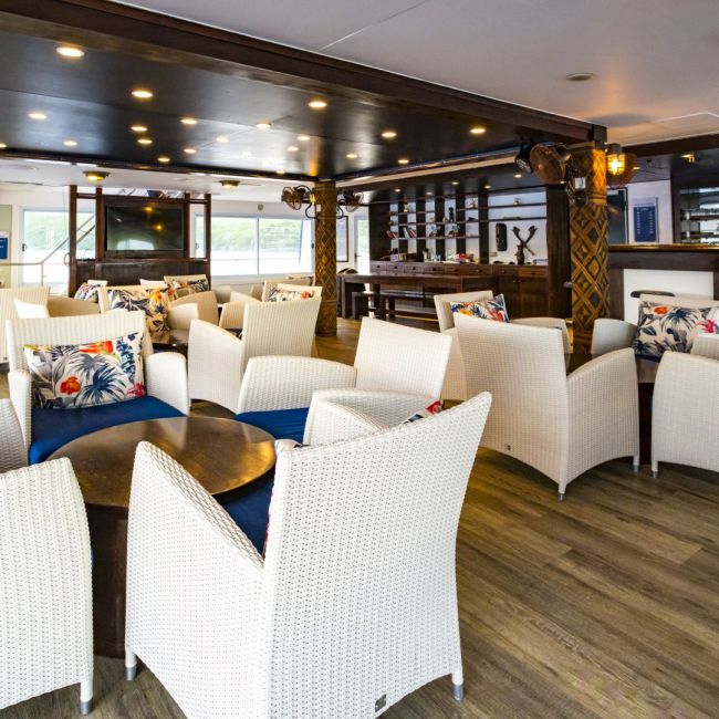 Luxury Cruise Ship Lounge and Bar Area An elegant indoor lounge and bar area aboard a luxury cruise ship, featuring cream-colored wicker armchairs arranged throughout the space, dark wood ceiling with ambient lighting, colorful tropical patterned pillows, and a sophisticated nautical-inspired design with wooden beams and decorative elements.