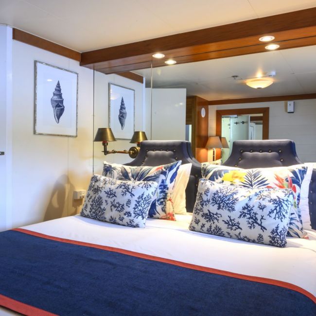 Luxury Cruise Ship Stateroom with Ocean View An elegantly designed cruise ship cabin featuring dual beds with navy upholstered headboards, nautical-themed decorative pillows, and a large mirror. The room showcases warm wood beam accents, recessed lighting, brass ship-style lamps, and coastal artwork, creating a sophisticated maritime atmosphere typical of modern luxury cruise accommodations.