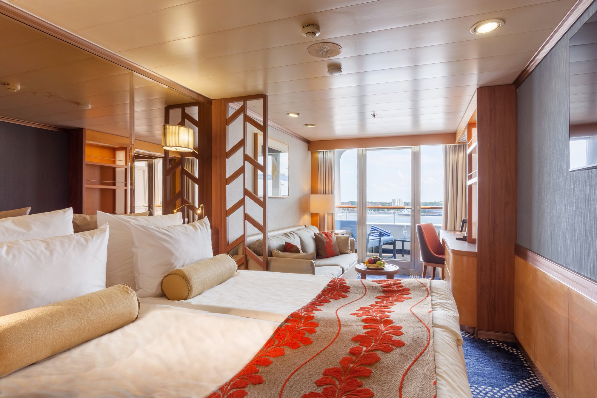 Luxury Cruise Ship Suite with Ocean View An elegant cruise ship cabin featuring a spacious bedroom with premium furnishings, wooden accents, and a separate living area with ocean view balcony overlooking the sea. The suite exemplifies modern cruise ship luxury accommodations with warm earth tones and contemporary design elements.