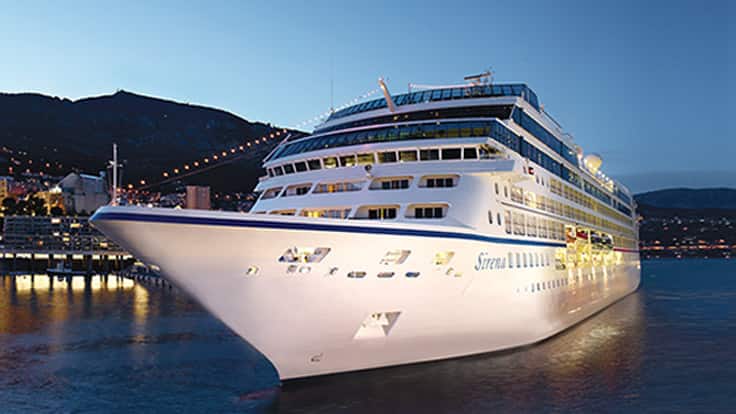 Luxury Cruise Ship at Night A pristine white cruise ship docked at a scenic harbor with mountains in the background during twilight hours. The vessel is illuminated, showcasing its elegant design against the dark blue evening sky.