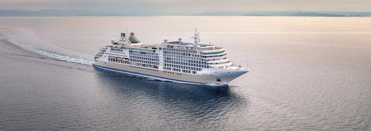A large, white luxury cruise ship sailing across calm, blue waters with a misty horizon in the background. The vessel appears to be a high-end cruise liner with multiple decks and modern design.