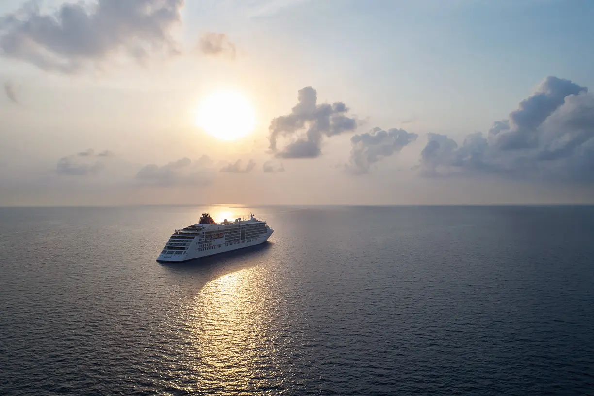 A large modern cruise ship floats peacefully on calm blue ocean waters during golden hour, with the sun rising or setting on the horizon and dramatic white clouds illuminating the sky. The vessel's multiple decks and sleek white hull are captured in perfect detail against the serene seascape.