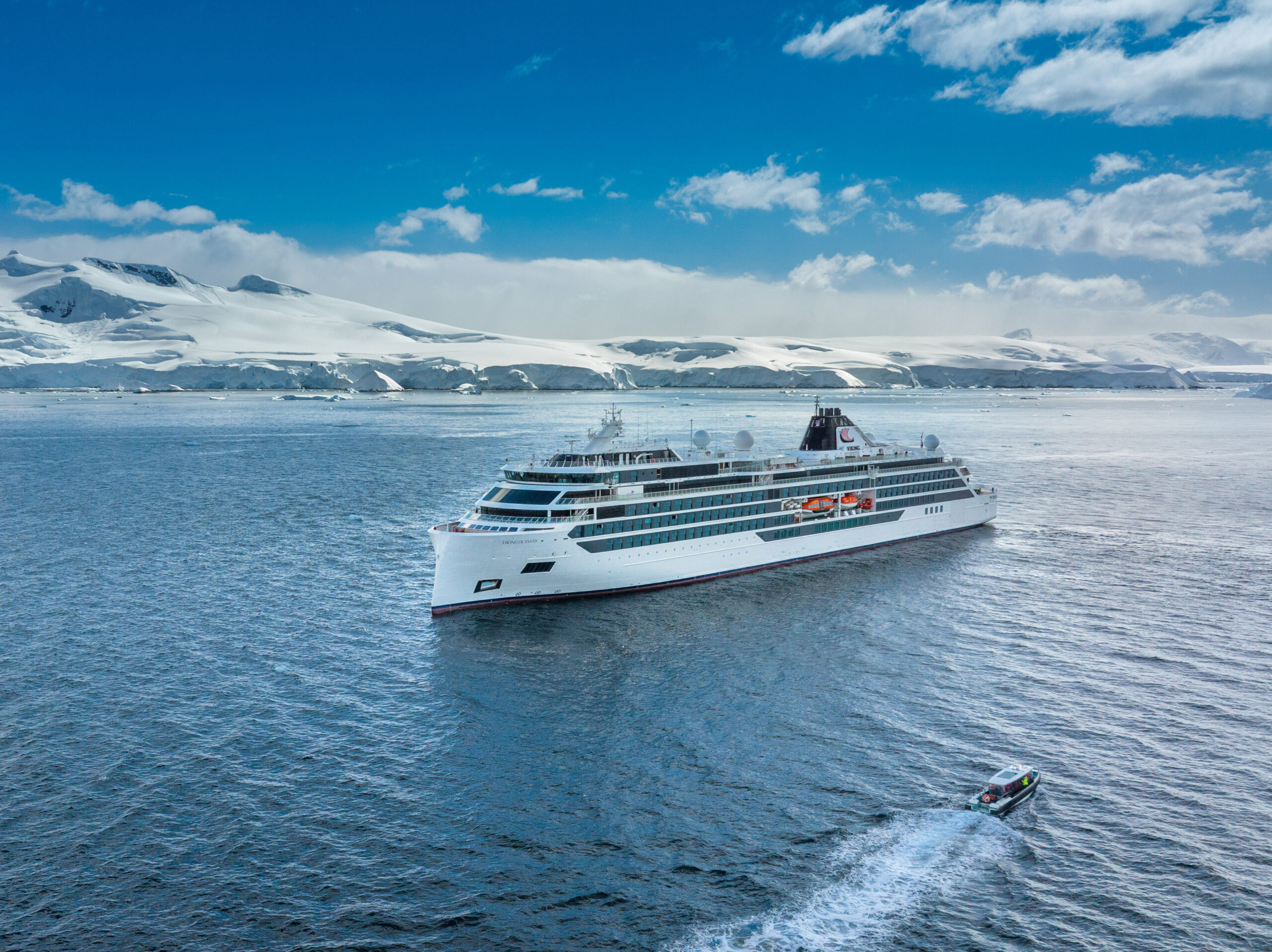 Luxury Cruise Ship in Antarctic Waters A pristine white luxury cruise ship navigating through icy blue waters with snow-covered mountainous Antarctic landscape in the background. A small support boat trails behind the vessel.