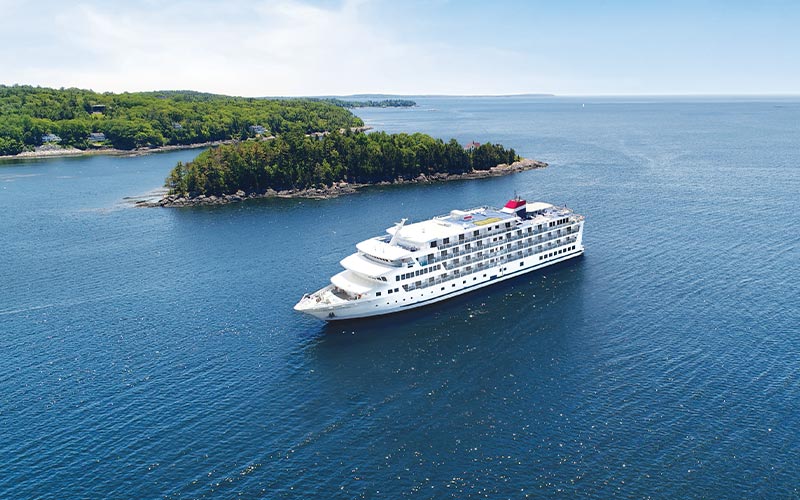 Luxury Cruise Ship in Coastal Landscape A white cruise ship sailing near a forested coastline with rocky shores and green islands. The vessel appears to be a small to mid-size luxury cruise ship navigating through calm blue waters.