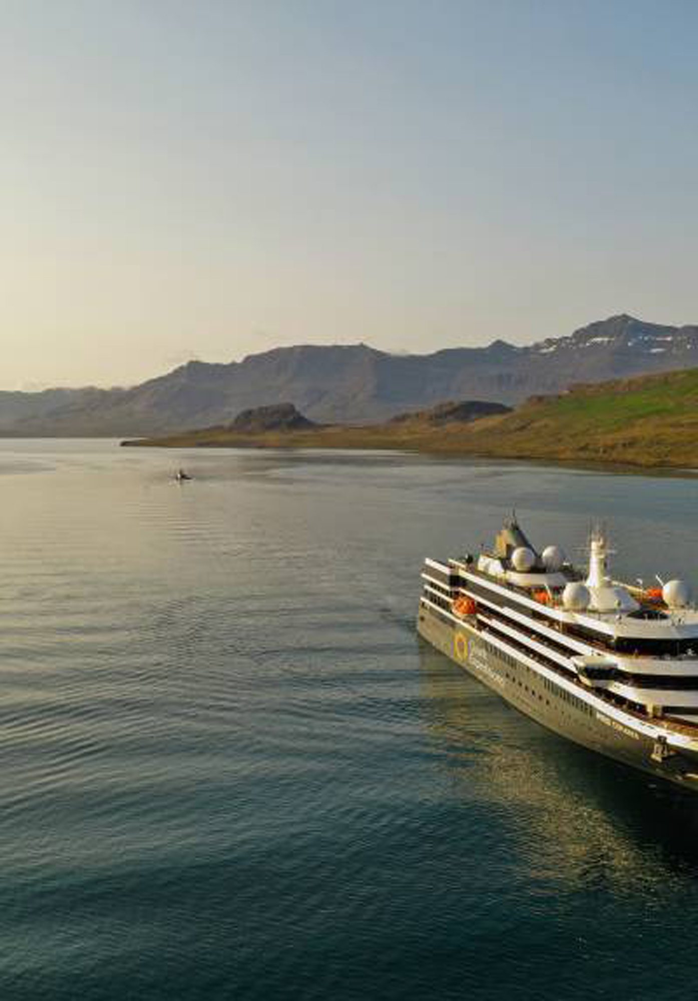 Luxury Cruise Ship in Mountainous Landscape A sleek, modern cruise ship navigates through calm waters with rugged mountain terrain in the background. The vessel is adorned with multiple satellite domes and has a clean, contemporary design.