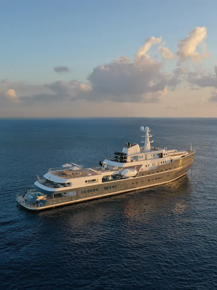 Luxury Expedition Yacht in Open Ocean A stunning golden-hulled expedition yacht anchored in calm blue waters during golden hour, with elegant multi-deck architecture featuring observation areas and a helicopter pad. The vessel is framed against a picturesque sky with soft cumulus clouds, exemplifying ultra-luxury small-ship cruising.