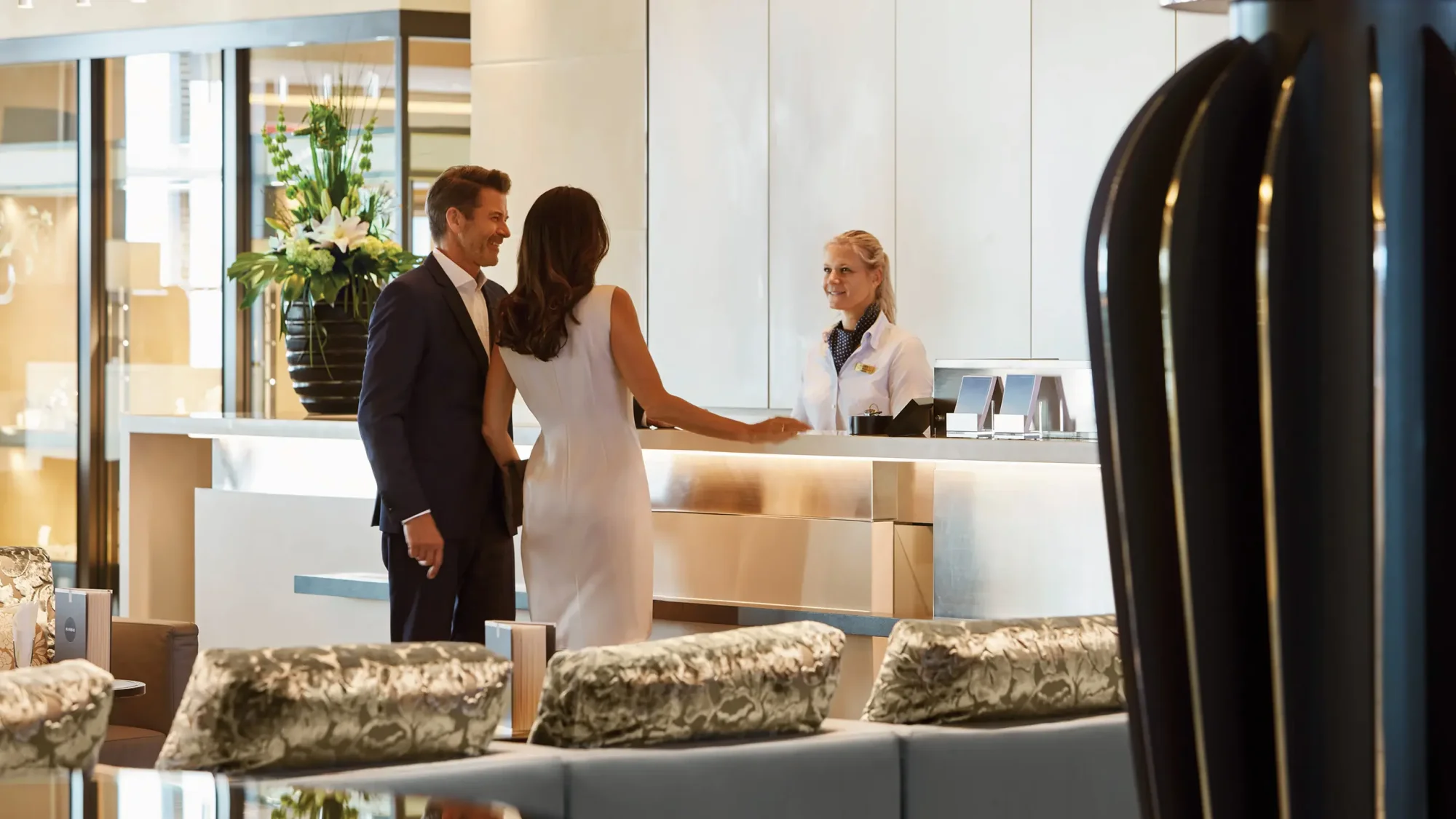 Luxury Hotel Reception Desk Check-in A sophisticated hotel lobby scene showing a well-dressed couple checking in at a modern reception desk with a blonde female staff member. The contemporary space features clean white panels, elegant furnishings with patterned cushions, decorative plants, and warm ambient lighting characteristic of upscale hospitality.