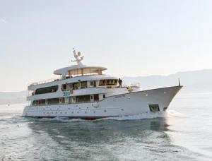 Luxury Motor Yacht Anchored in Calm Waters A sophisticated multi-deck motor yacht with golden-toned wooden upper decks and white hull anchors in calm, shallow turquoise waters with mountainous coastline in the misty background. The vessel features multiple levels with a distinctive bridge and appears to be a high-end expedition or cruise yacht designed for luxury travel and exploration.