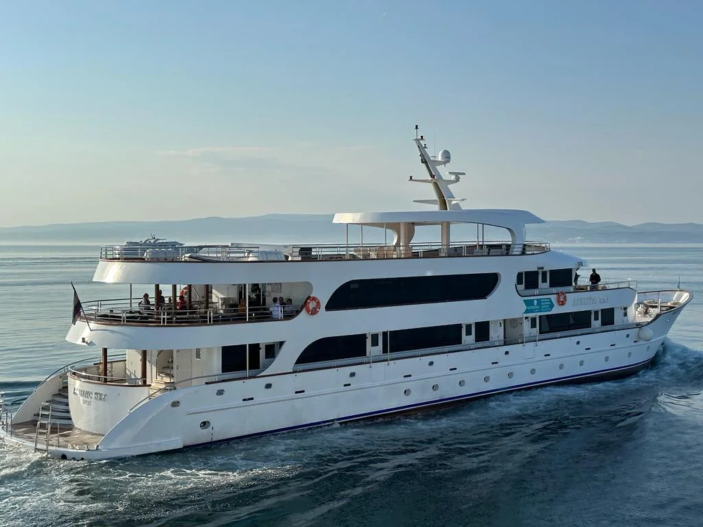 Luxury Motor Yacht Anchored in Mediterranean Waters A striking white multi-deck motor yacht with sleek modern design anchored in calm blue Mediterranean waters, featuring multiple passenger decks, outdoor terraces, and sophisticated navigation equipment, with mountainous coastline visible in the distance.