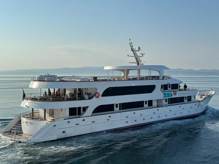 Luxury Motor Yacht in Calm Waters A elegant white motor yacht with multiple decks and modern amenities cruises through calm, clear blue waters with mountainous coastal terrain visible in the background. The vessel features contemporary design with dark accents, outdoor deck spaces, and sophisticated navigation equipment on the bridge.