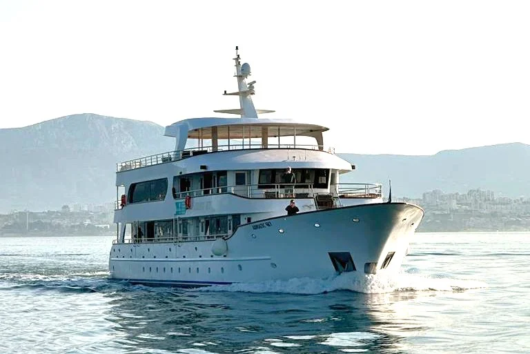 Luxury Motor Yacht in Coastal Waters A sophisticated multi-deck motor yacht with light blue and white hull cruising through calm coastal waters with mountainous terrain visible in the background. The vessel features modern design elements including an upper observation deck, multiple levels, and professional navigation equipment.