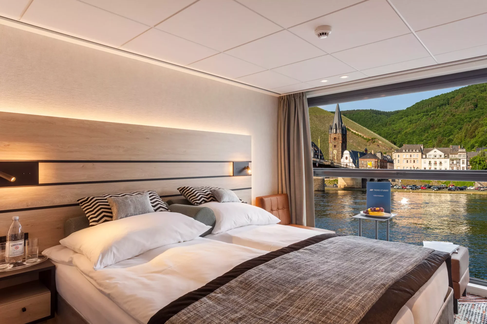Luxury River Cruise Cabin with Rhine Valley View A sophisticated river cruise cabin featuring a modern bedroom with panoramic floor-to-ceiling windows overlooking a picturesque Rhine Valley town with church steeple and vineyards. The contemporary design includes warm wood accents, ambient lighting, and elegant furnishings with a view of the river and charming European architecture.