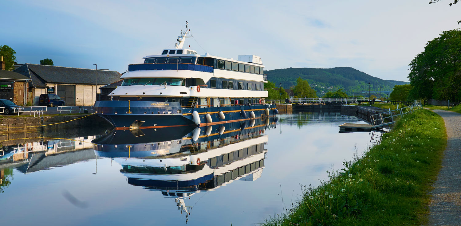 Luxury River Cruise Vessel at Dock A multi-deck luxury river cruise ship moored at a peaceful inland waterway, featuring distinctive blue and white exterior with white fenders along the hull. The vessel is docked alongside a charming riverside community with tree-lined banks and forested hills visible in the background.