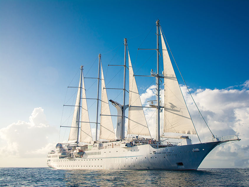 A magnificent white multi-masted sailing vessel cruising on bright blue waters with white sails fully extended against a clear sky with soft clouds.