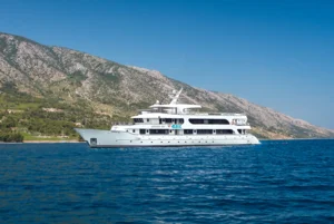 Luxury Yacht Anchored in Mediterranean Waters A pristine white luxury motor yacht anchored in crystal-clear turquoise Mediterranean waters with a dramatic rocky coastline and green hillside in the background, representing upscale cruising and Mediterranean destinations.