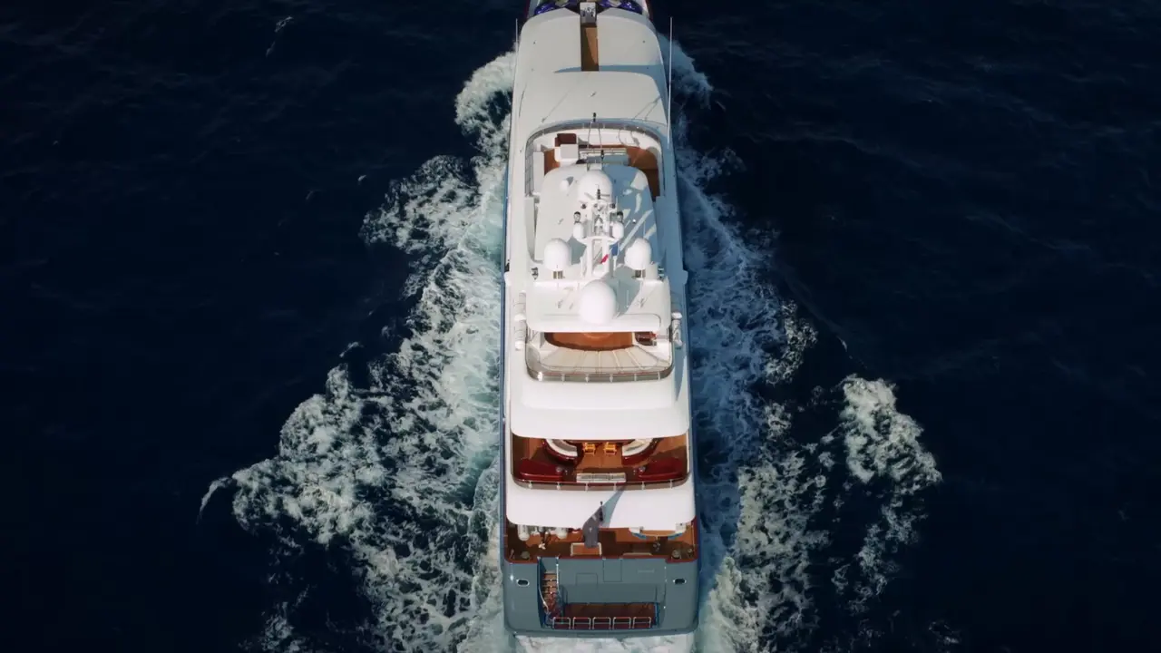 Luxury Yacht Cruising at Full Speed An overhead aerial view of a multi-deck white luxury motor yacht cutting through deep blue ocean waters, creating dramatic white wake patterns on both sides. The vessel displays multiple teak-accented deck levels and is captured at high speed during daytime.