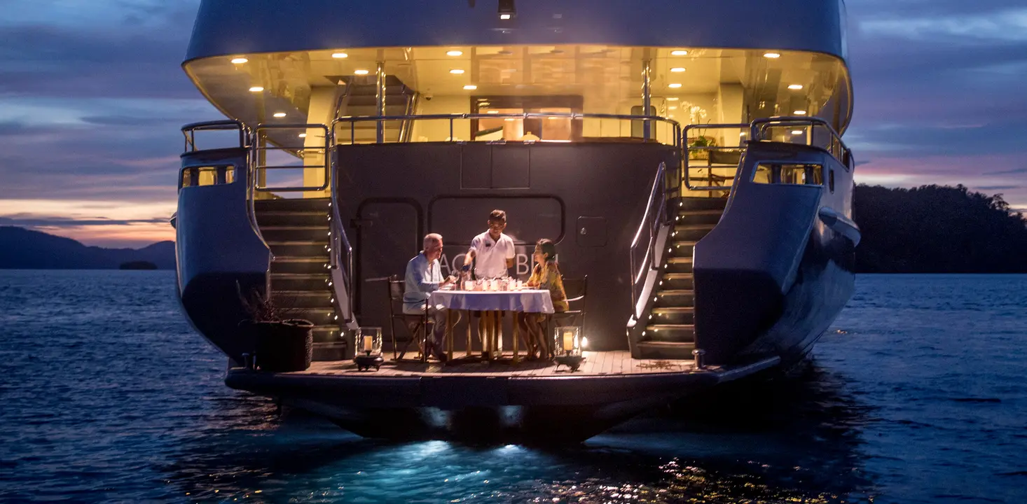 Luxury Yacht Stern Dining at Twilight An elegant outdoor dining experience on the illuminated stern platform of a luxury yacht during golden hour, with a family of four seated at a formally set table while the sun sets over calm coastal waters.