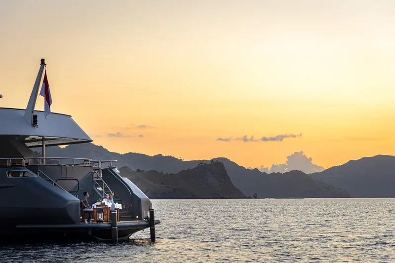 Luxury Yacht at Tropical Sunset A modern luxury yacht tender is moored at sunset with dramatic golden-orange skies reflecting off calm waters, with mountainous tropical islands silhouetted in the background. The scene captures the tranquil beauty of an upscale cruise or yacht experience in an idyllic island destination.