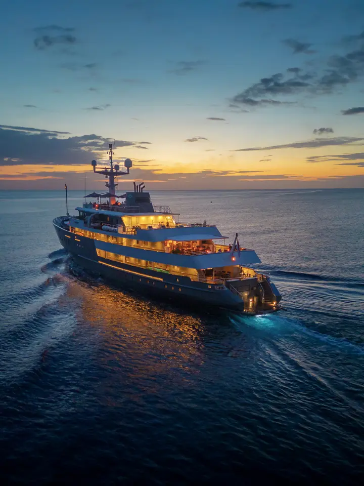 Luxury Yacht at Twilight with Golden Lights A stunning expedition yacht illuminated by warm golden interior lights anchors in calm waters during a vibrant sunset, with the vessel's navigation equipment visible against a dramatic sky transitioning from orange and yellow to deep blue.