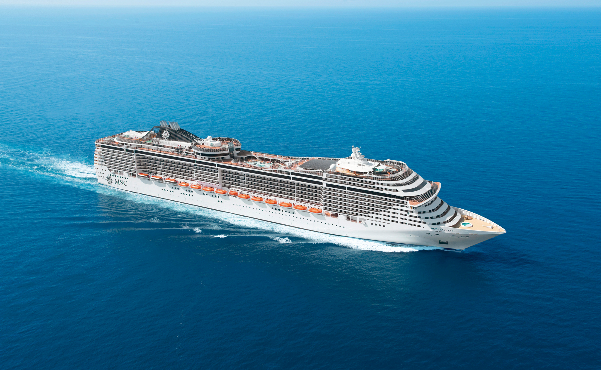 MSC Cruise Ship Sailing Open Ocean A large white and gray MSC cruise ship sailing through vibrant blue ocean waters, showcasing multiple decks and orange lifeboat details.