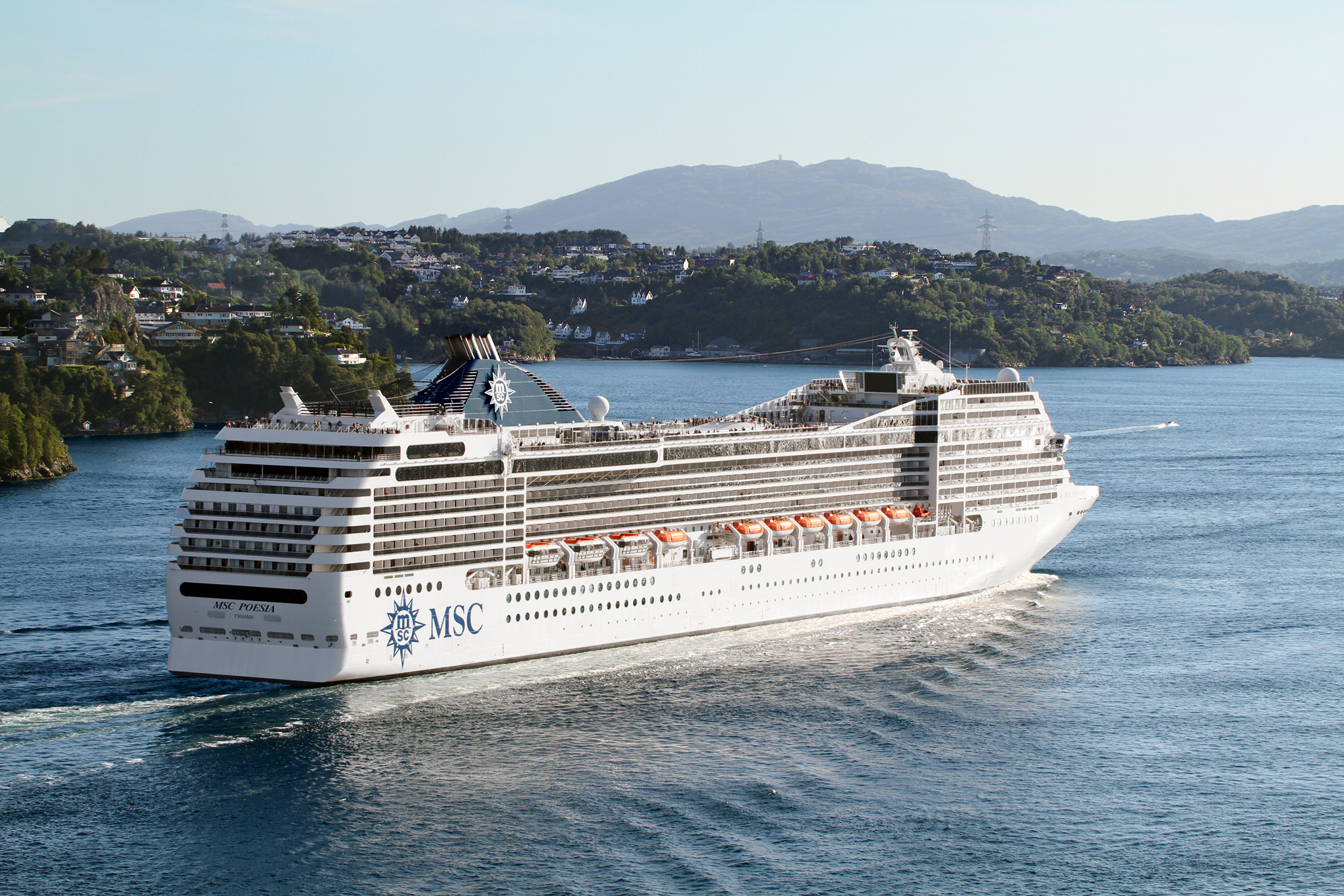 MSC Cruise Ship in Scenic Coastal Landscape A large white MSC cruise ship sailing through a picturesque bay with wooded shorelines and mountains in the background. The vessel is cruising on calm blue waters with a lush green landscape and residential area visible.