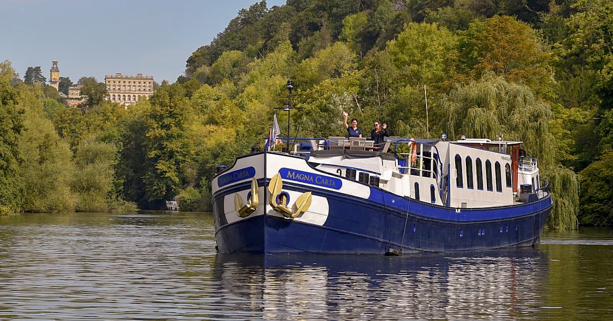 Magna Carta River Barge on European Waterway A luxury river cruise barge named Magna Carta navigates a picturesque European river surrounded by lush forests and historic architecture, with a grand palazzo-style building visible on the hillside in the background.