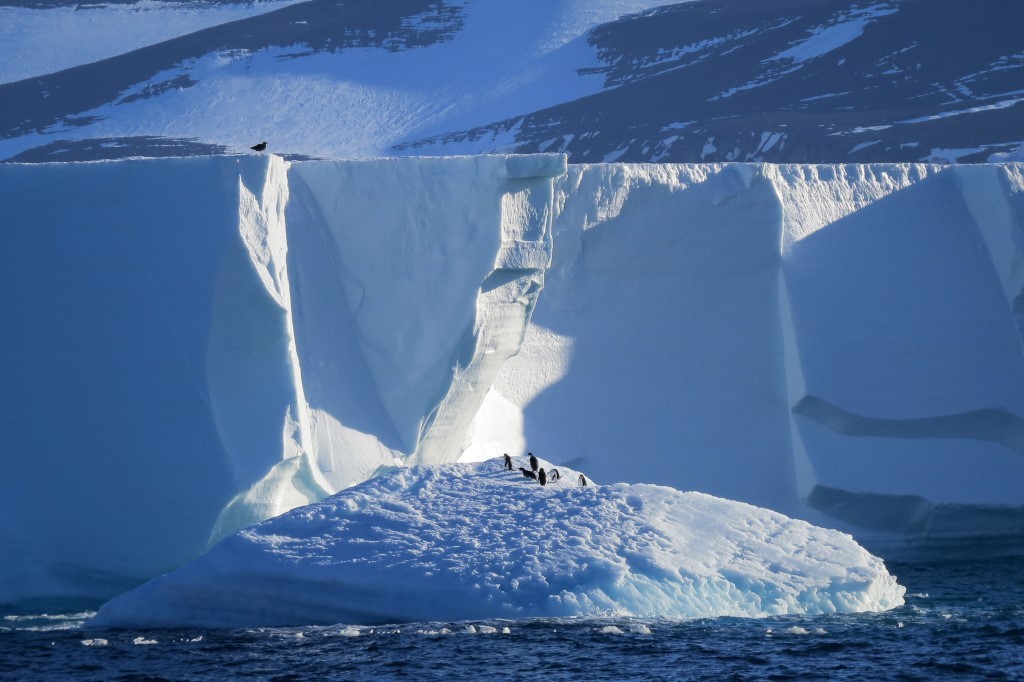Majestic Iceberg in Antarctic Waters A stunning massive iceberg with towering white walls and deep blue crevasses rises dramatically from the Antarctic Ocean, with a small group of explorers visible on its peak and snow-covered mountains visible in the background, showcasing the raw beauty and scale of polar exploration.