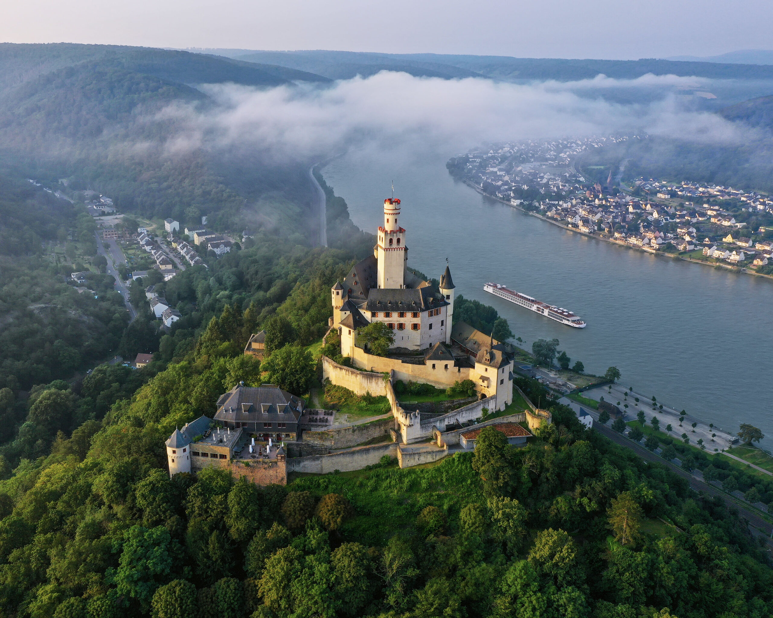 Marksburg Castle on the Rhine River An aerial view of the iconic Neuschwanstein Castle perched on a hilltop above the Rhine River in Germany, with a river cruise ship navigating the misty valley below. The castle's distinctive yellow limestone walls and towers rise majestically from dense forests, while morning fog blankets the picturesque riverside towns and vineyards along the river bend.