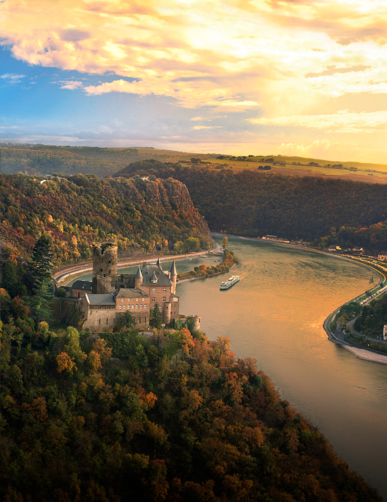 Medieval Castle on Rhine River Bend A historic stone castle perched on a forested riverbank with a dramatic curved river and golden sunset landscape. A river cruise ship navigates the scenic waterway past the picturesque medieval fortress.