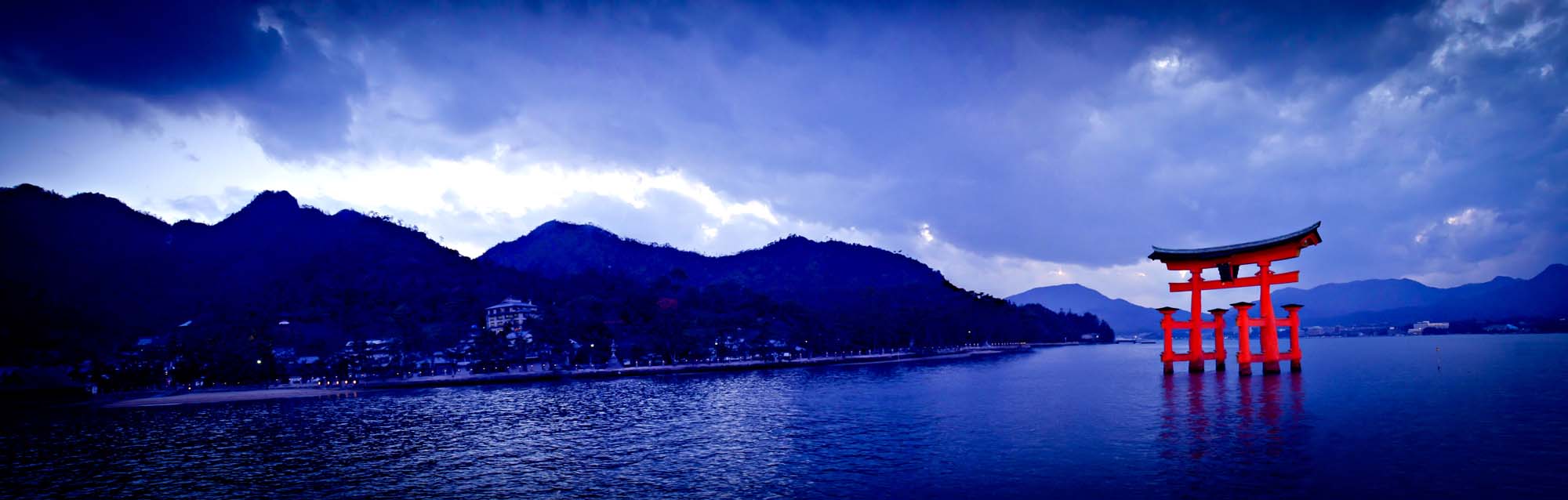 Miyajima’s Floating Torii Gate at Twilight A vibrant red torii gate stands in the blue waters of Miyajima, with misty mountains and a coastline in the background during a serene twilight hour.
