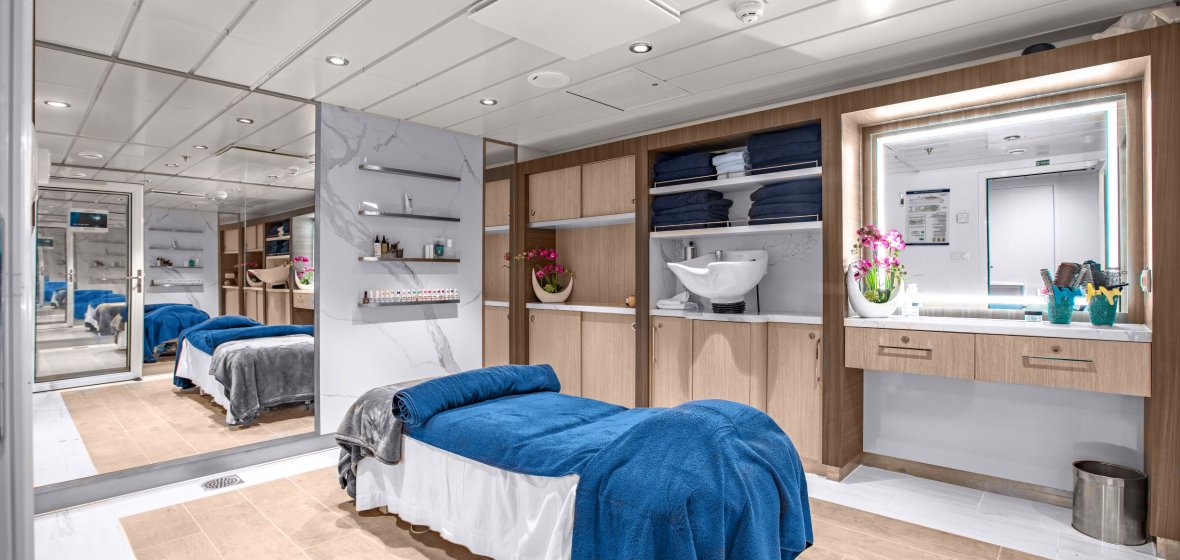 A luxurious and contemporary cruise ship cabin featuring a spacious bedroom with blue bedding, integrated marble bathroom with dual sink vanity, and clever built-in storage solutions. The suite showcases modern design elements including wood cabinetry, recessed lighting, and mirrored closets that enhance the sense of space in this well-appointed ocean-going accommodation.