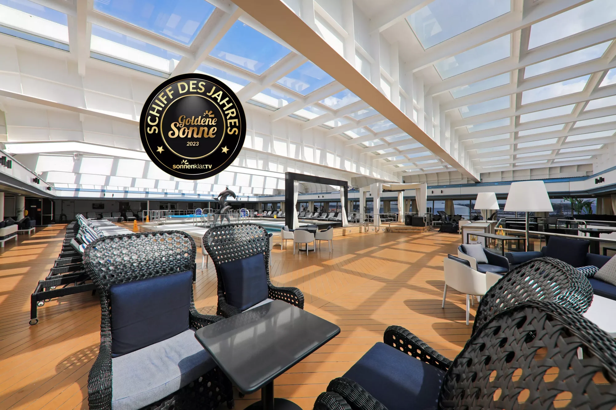 Modern Indoor Pool Deck with Skylight Architecture A contemporary indoor aquatic complex featuring a large swimming pool, modern black mesh loungers with blue cushions, and an expansive skylight ceiling that floods the space with natural sunlight. The sophisticated design includes wooden flooring, casual seating areas, and panoramic windows overlooking outdoor facilities, earning recognition as the Ship of the Year (Schiff des Jahres) in 2023.