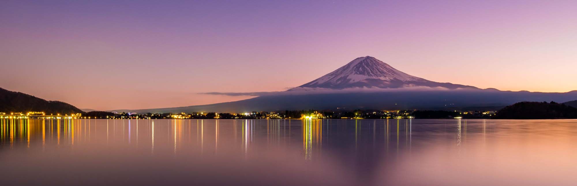 Mount Fuji at Sunset Over Lake Kawaguchiko A stunning panoramic view of the iconic Mount Fuji reflecting in calm waters at twilight, with city lights illuminating the shoreline in soft golden and purple hues.