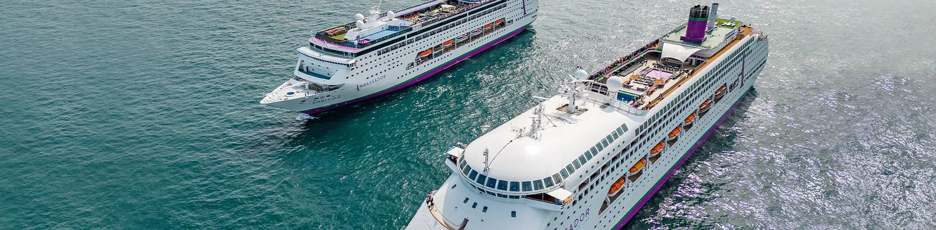 A large modern cruise ship named Ambassador, featuring white hull with distinctive purple and orange accent bands, docked at a tropical port with clear blue waters. The vessel displays multiple decks, communication satellites, and lifeboats typical of contemporary ocean liner design.