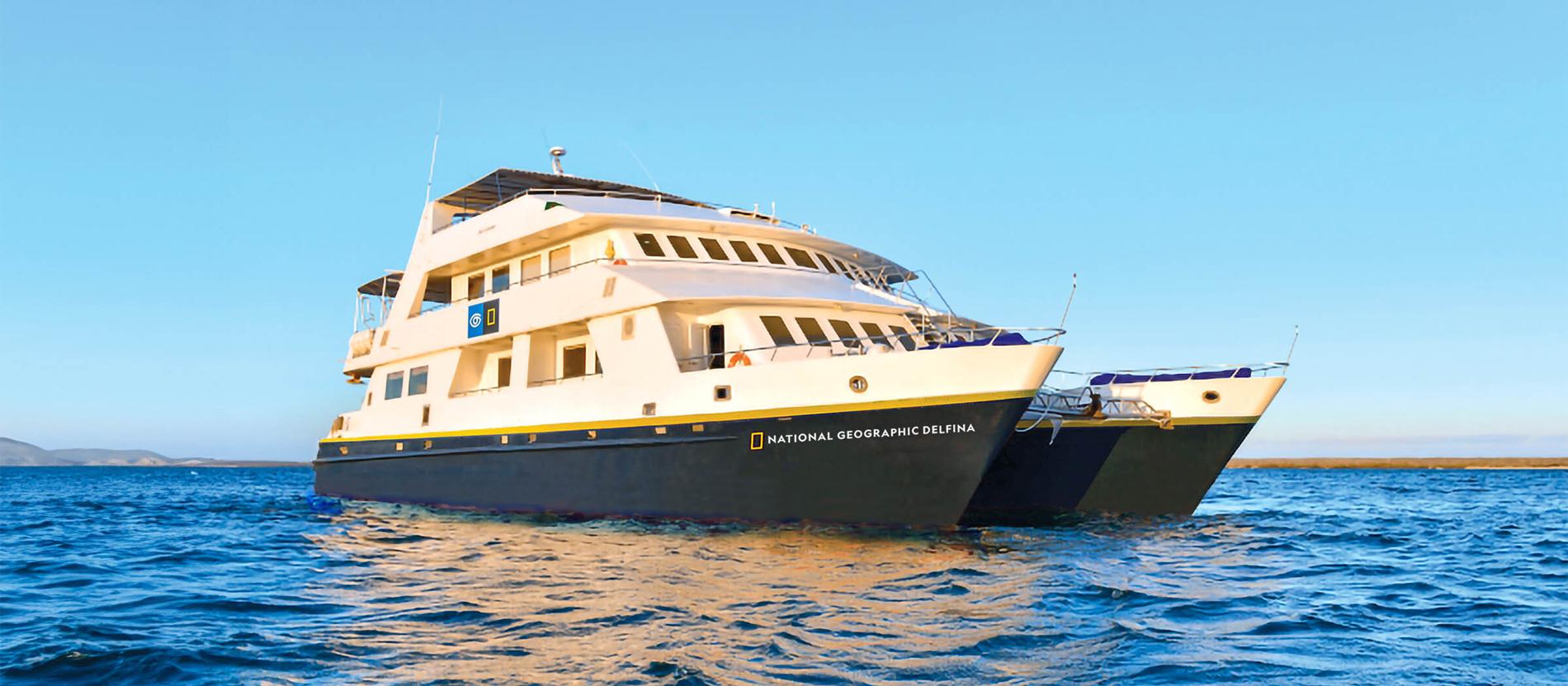National Geographic Delfina Cruise Ship at Sea The National Geographic Delfina, a sophisticated expedition cruise vessel, anchors in crystalline turquoise waters under clear blue skies. The multi-deck ship features cream and navy hull coloring with characteristic National Geographic branding, representing premium small-ship cruise experiences in tropical destinations.
