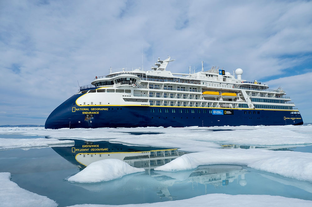National Geographic Endurance in Arctic Ice The National Geographic Endurance expedition cruise ship navigates through Arctic sea ice, with its distinctive dark blue and yellow hull reflected in the calm, icy waters surrounding it. The vessel is specifically designed for polar exploration and environmental research in extreme conditions.