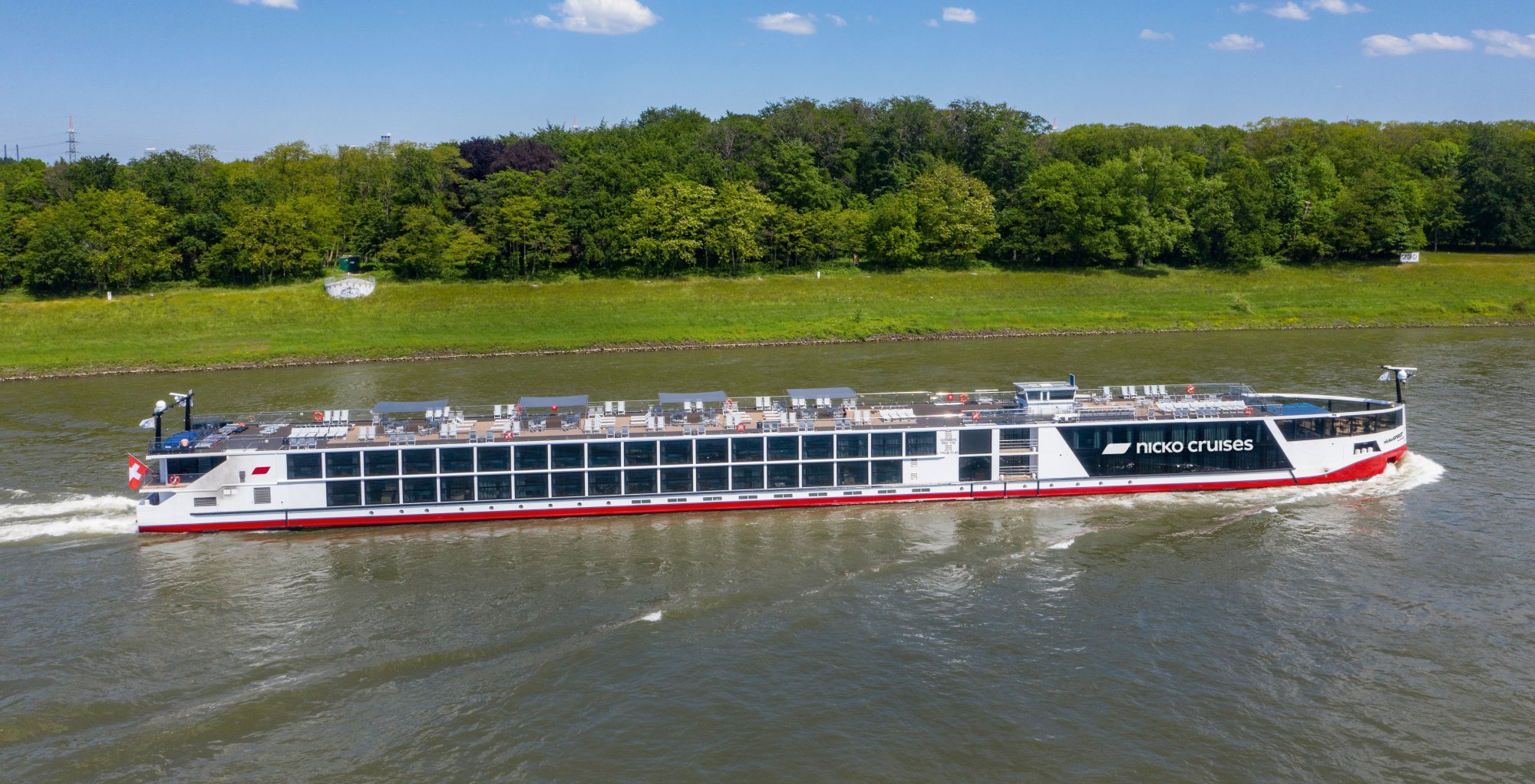 Nicko Cruises River Vessel on European Waterway A modern Nicko Cruises river cruise ship navigates a European river with lush green countryside visible along the banks. The vessel features extensive deck cargo and continuous window lines characteristic of contemporary river cruise design.