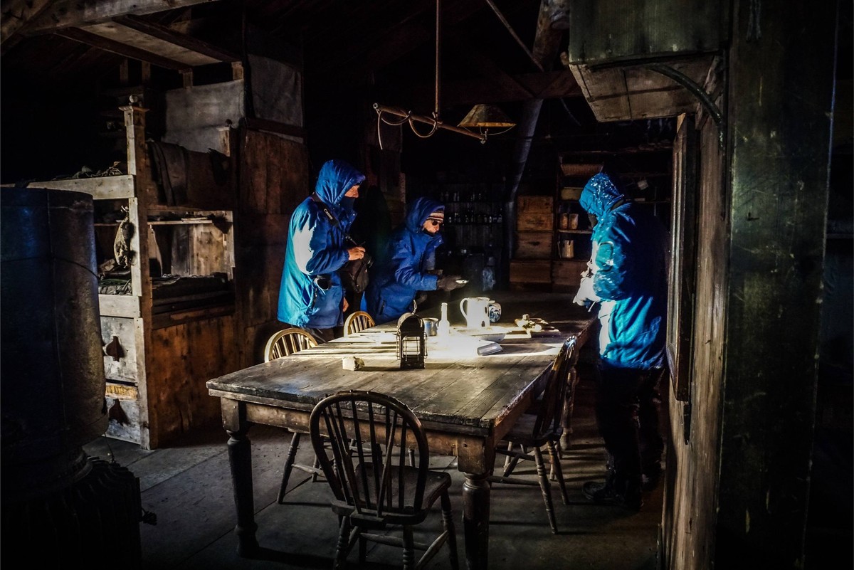 Night Gathering in Historic Industrial Space Three people in bright blue jackets gather around a weathered wooden table in a dimly-lit, rustic industrial setting with exposed brick and metal structures. The atmospheric nighttime scene captures an intimate moment in what appears to be a historic warehouse or ship-related facility, illuminated by natural light creating strong contrast with the dark surroundings.