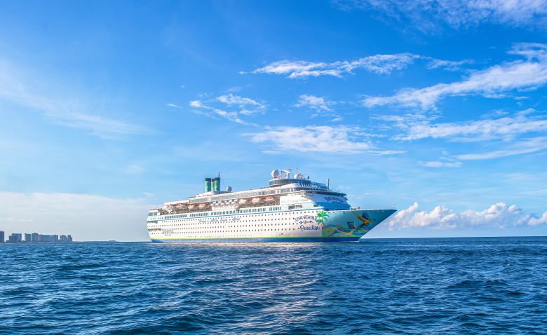 A large Norwegian Cruise Line ship cruising across bright blue ocean waters with a cloudy sky in the background. The ship features the distinctive blue and white color scheme of the cruise line.