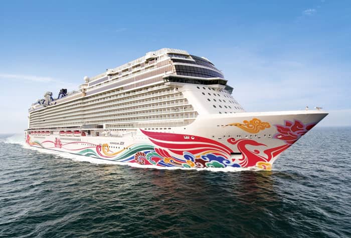 A large, colorful Norwegian cruise ship sailing on open water, featuring a vibrant red, yellow, and blue hull design. The ship appears modern and sleek against the blue sky and ocean.