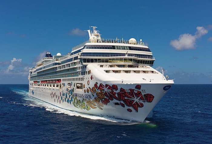 A colorful Norwegian Cruise Line ship sailing across a bright blue ocean with a clear sky and white clouds in the background. The ship features a distinctive artistic hull design with colorful graphics.