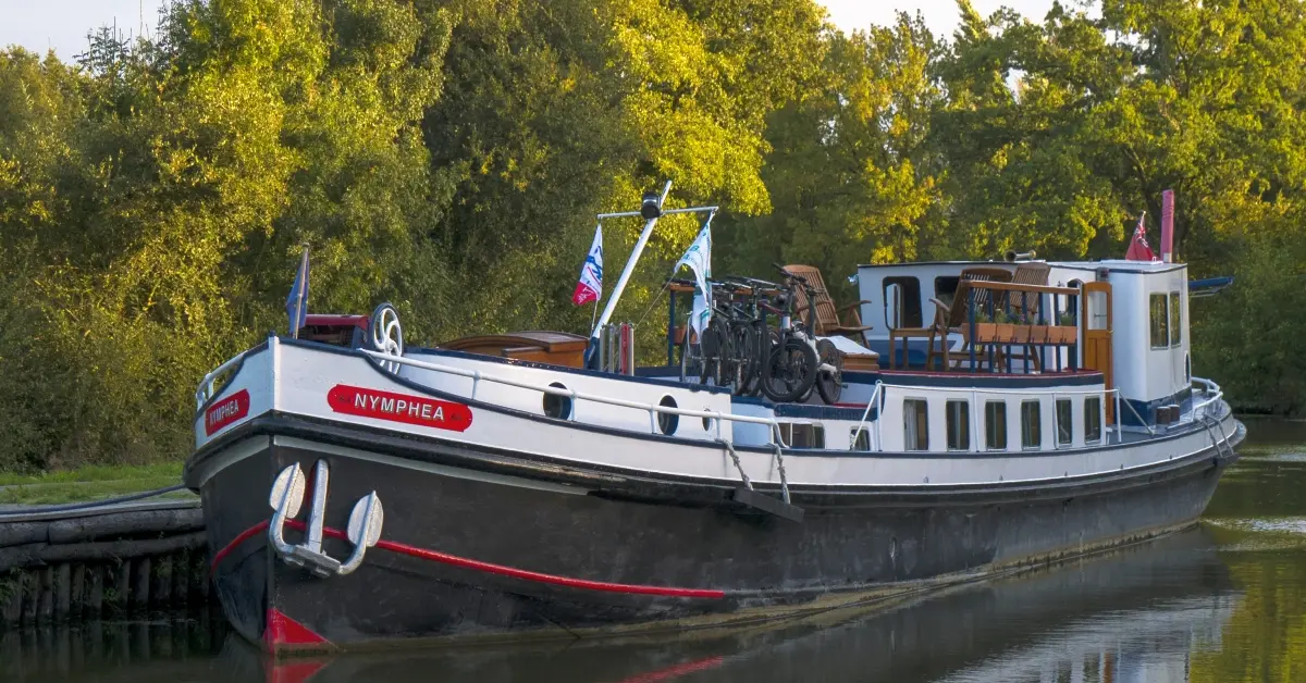 Nymphea Barge Moored Along European Waterway A charming traditional European barge named Nymphea is moored along a peaceful riverside, featuring classic red and white exterior styling with multiple decks, cabin windows, and period bicycles displayed on deck. The vessel is surrounded by lush green foliage and willows, exemplifying leisurely river cruise tourism.