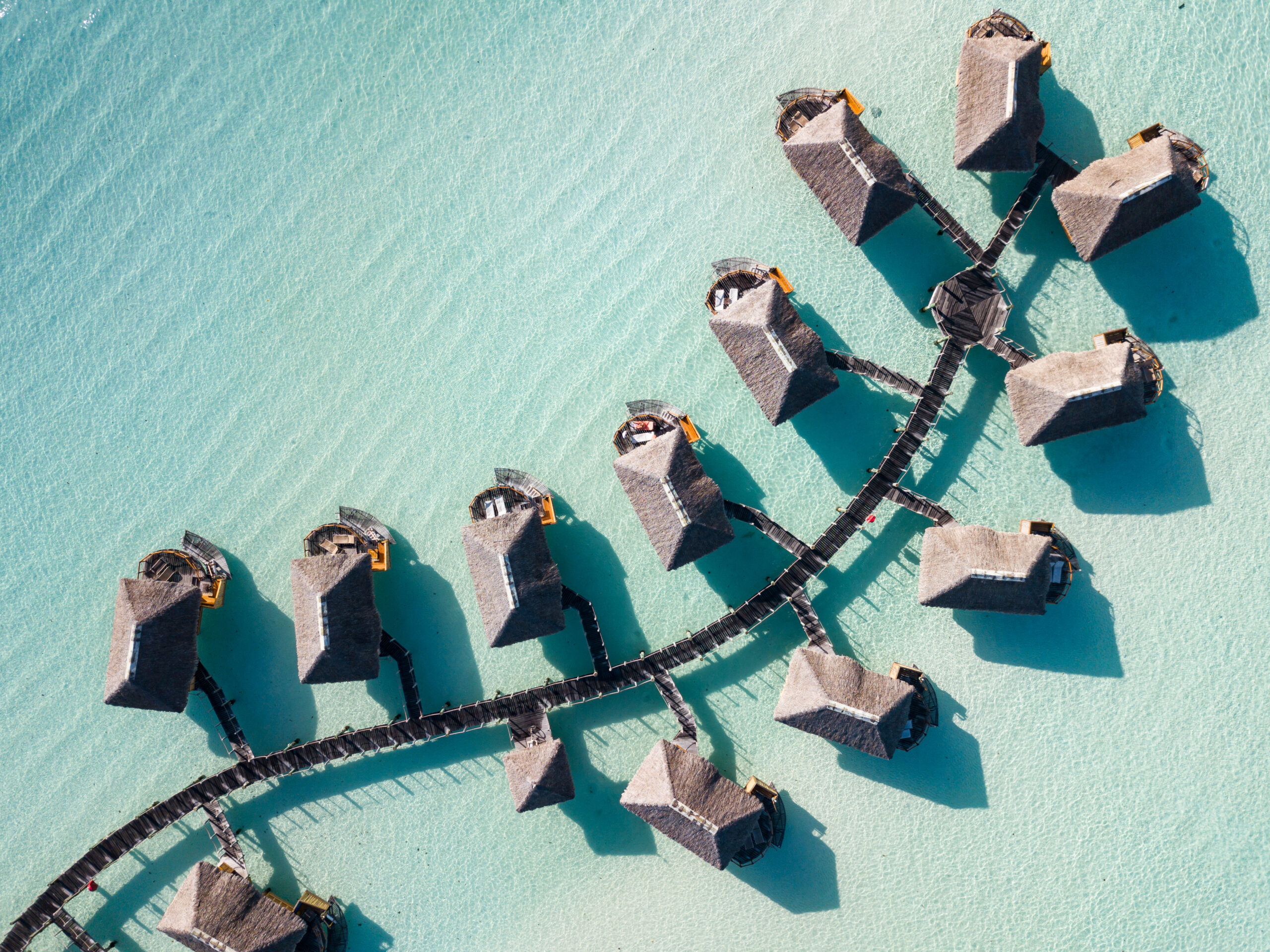 An aerial view of a luxury resort with thatched-roof overwater bungalows arranged in a curved pattern along wooden walkways in crystal-clear turquoise lagoon waters. The image showcases the pristine tropical destination with perfect symmetry and shallow waters characteristic of South Pacific island resorts.