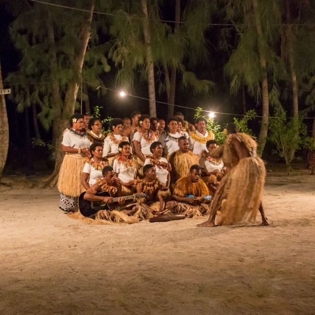 Pacific Island Cultural Evening Performance A vibrant nighttime cultural gathering on a sandy beach featuring a large group of performers in traditional Pacific Islander attire, including grass skirts and flower leis, arranged in a circle under string lights and towering palm trees. The scene captures an authentic ceremonial or entertainment experience typical of island excursions offered by cruise lines visiting the South Pacific.