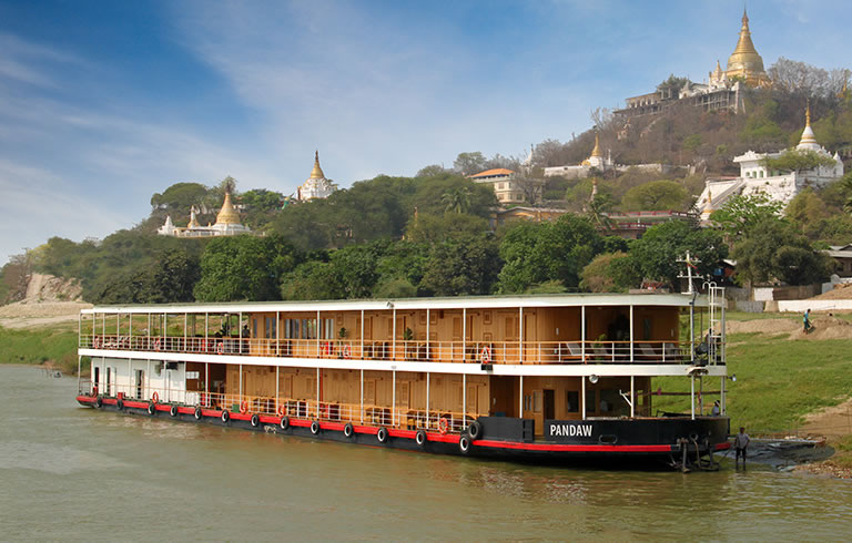 Pandaw River Cruise in Myanmar A multi-deck river cruise boat named Pandaw navigating a river in Myanmar with golden Buddhist pagodas and green hillsides in the background.