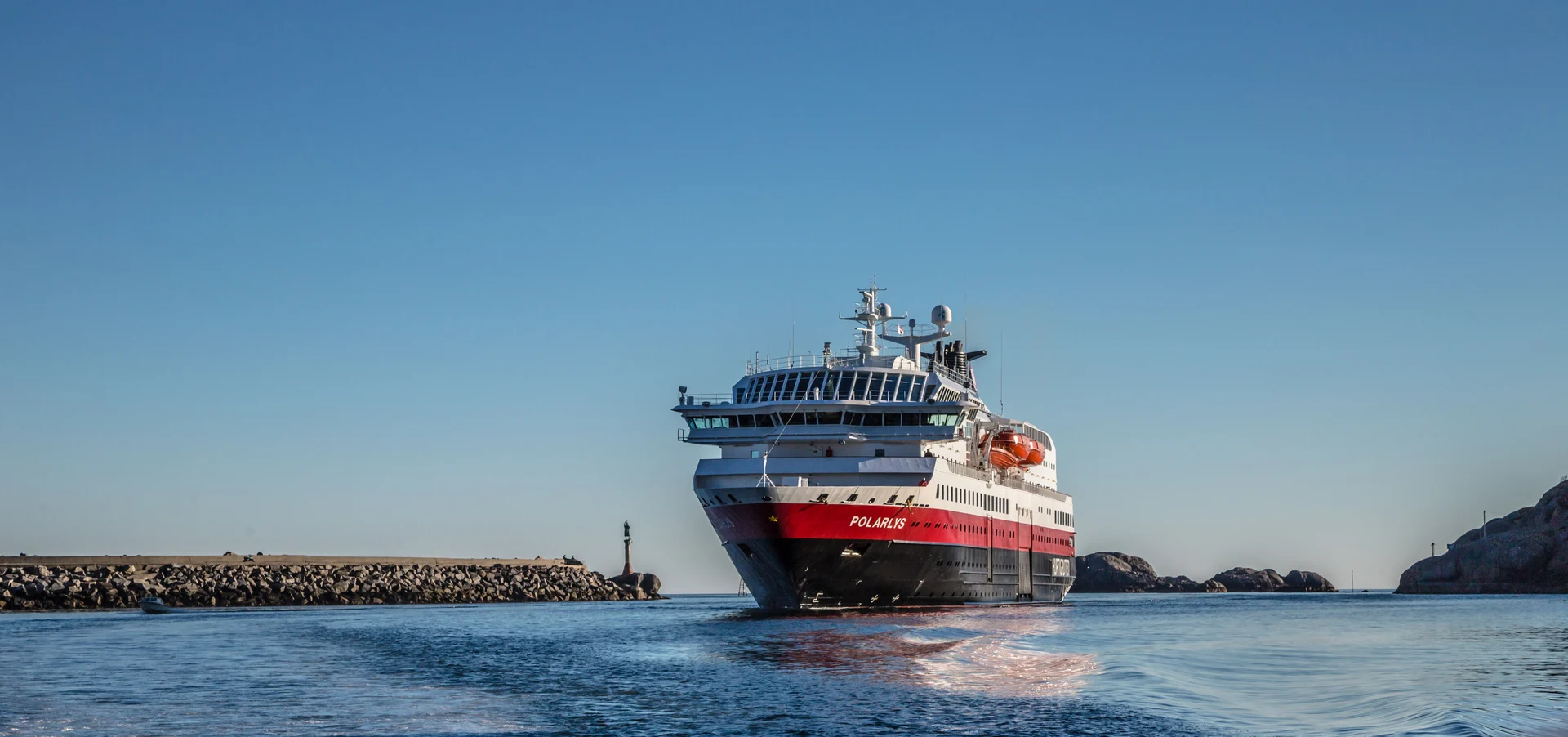 A modern cruise ship with red and white livery named Polaris navigating through a rocky coastal harbor with calm blue waters and clear sky.