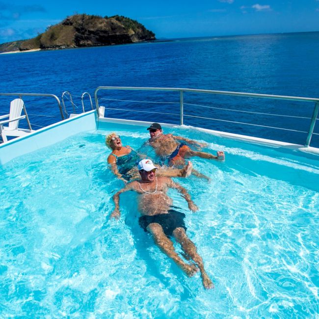 Poolside Tropical Paradise on Cruise Ship A family enjoys swimming and relaxing in a turquoise pool deck aboard a cruise ship, with pristine tropical waters and a lush island visible in the background under clear blue skies.