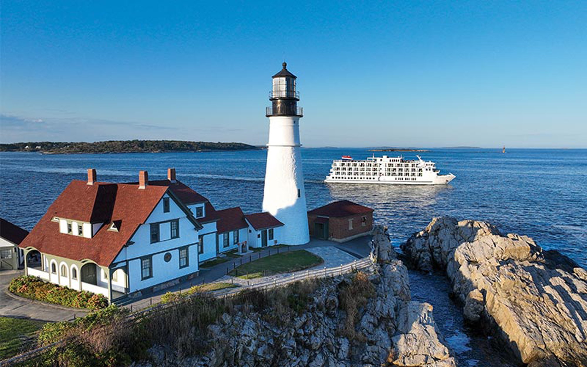 A scenic coastal view of the iconic Portland Head Light lighthouse with its distinctive white tower and black top, accompanied by a charming blue keeper's house and supporting buildings perched on rocky cliffs. A modern cruise ship can be seen anchored in the calm blue waters offshore, showcasing this popular New England cruise destination.