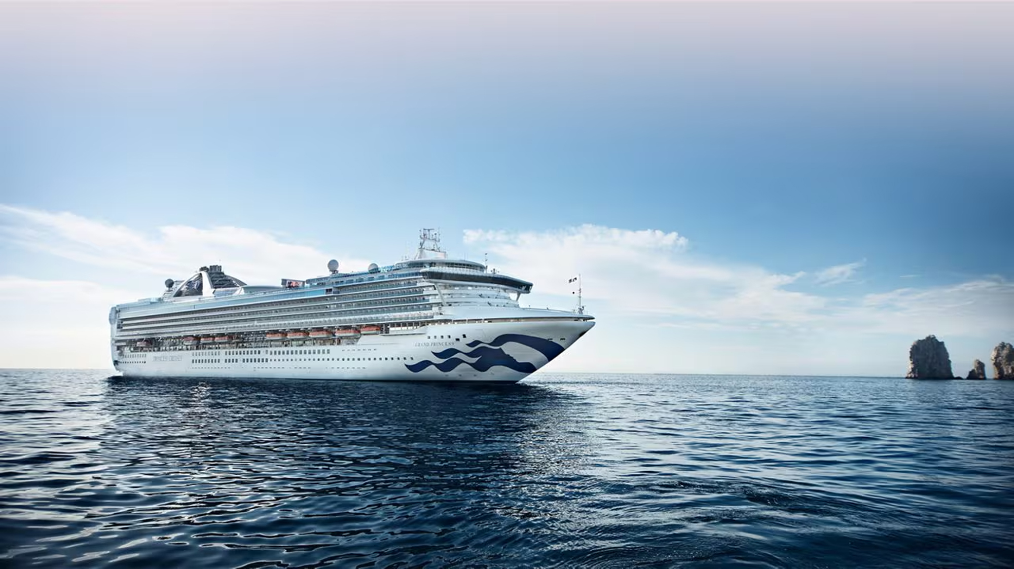 A sleek white and blue Princess cruise ship sailing on calm blue waters with rocky coastal formations in the background under a clear sky.